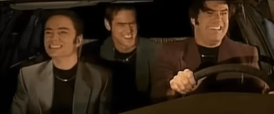 The image shows three men in a car, with the driver in the front and two passengers in the back. The driver is smiling…