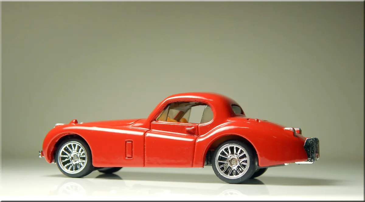 A red die-cast model of a classic two-door sports car is shown in profile against a plain gray background. The car has…