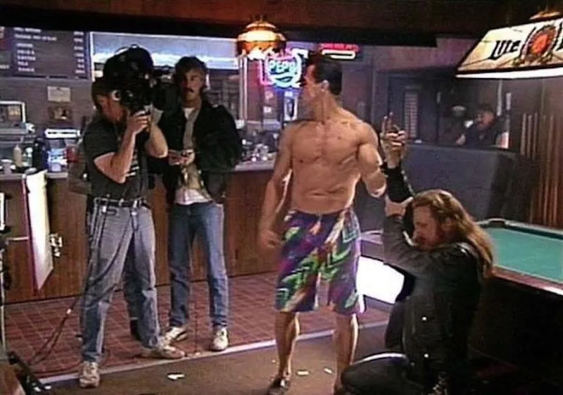 A shirtless man in colorful swim trunks stands in a bar, gesturing with his hands while a man with long hair kneels beside…