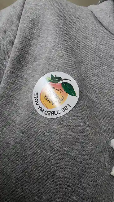 A circular sticker with a peach illustration and green leaves is stuck on a gray hoodie. The text on the sticker reads 'I…