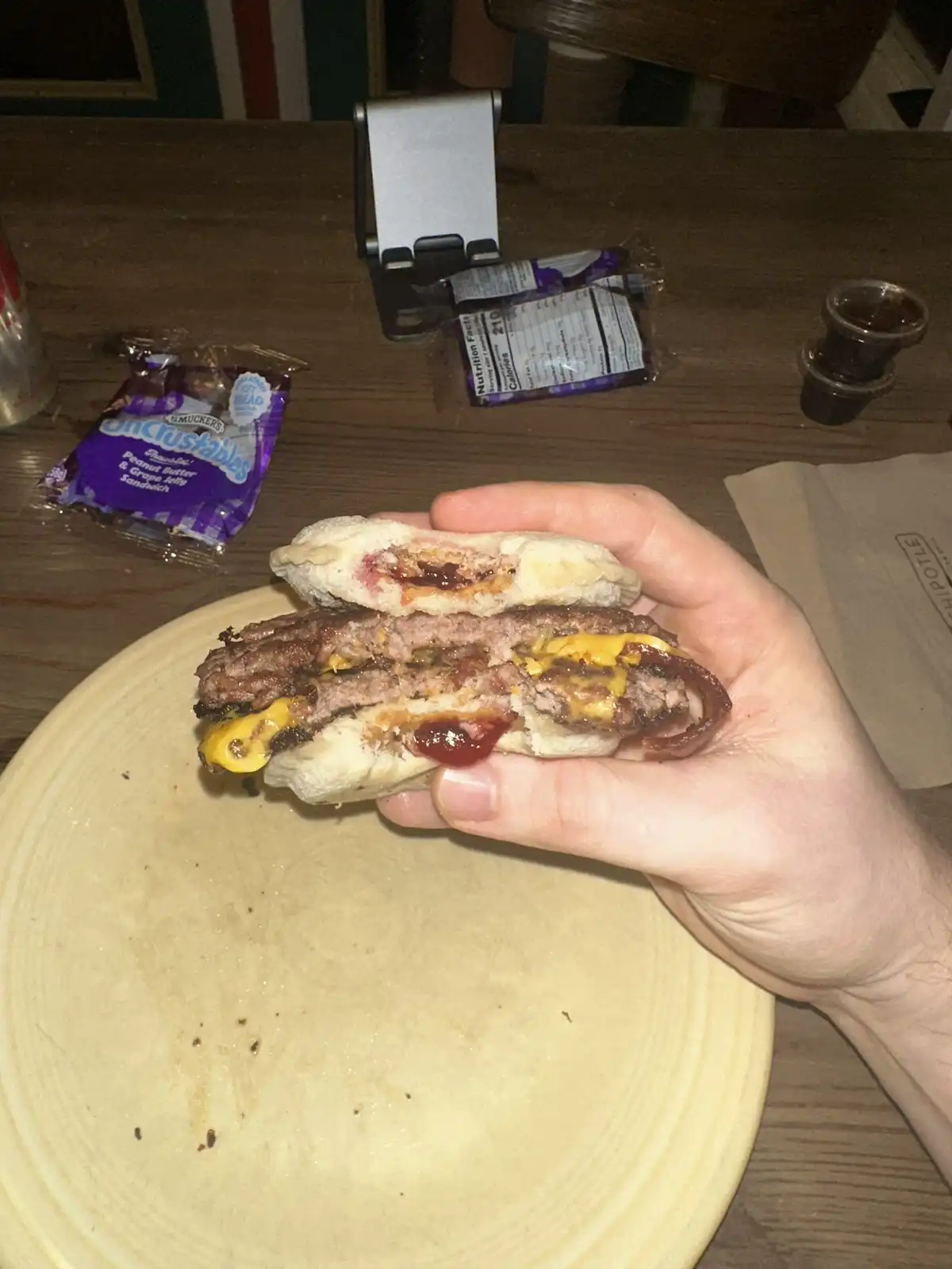 A hand holds a burger that has been sliced open, revealing a peanut butter and grape jelly sandwich inside the bun. In the…