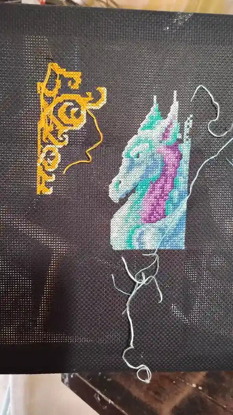 A close-up of a cross-stitch project on dark fabric shows a partially stitched unicorn head with a purple mane and teal…