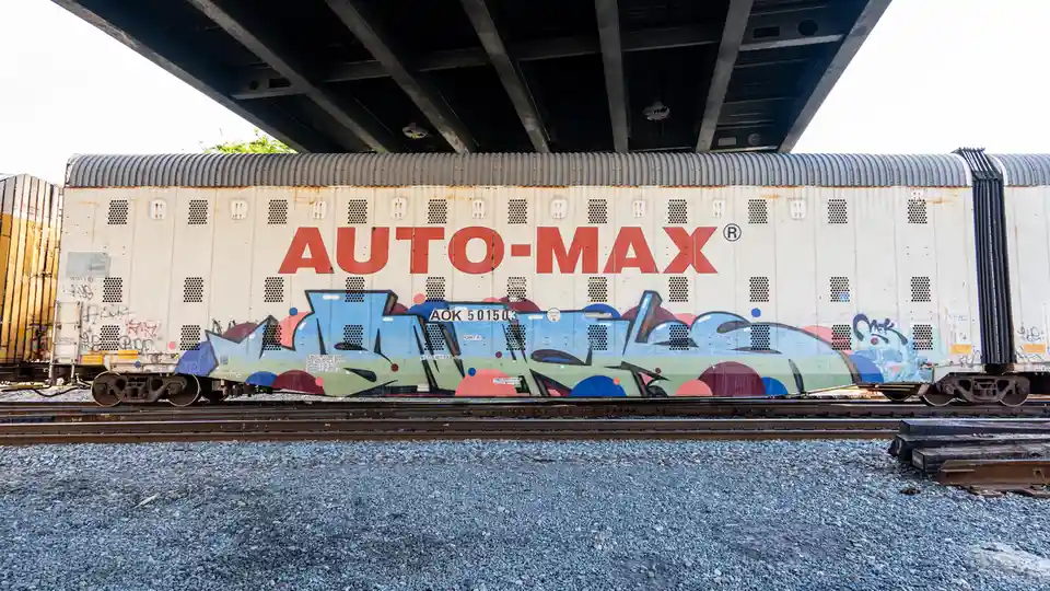 A white freight train car with 'AUTO-MAX' in large red letters is parked under a bridge, covered in colorful graffiti. The…