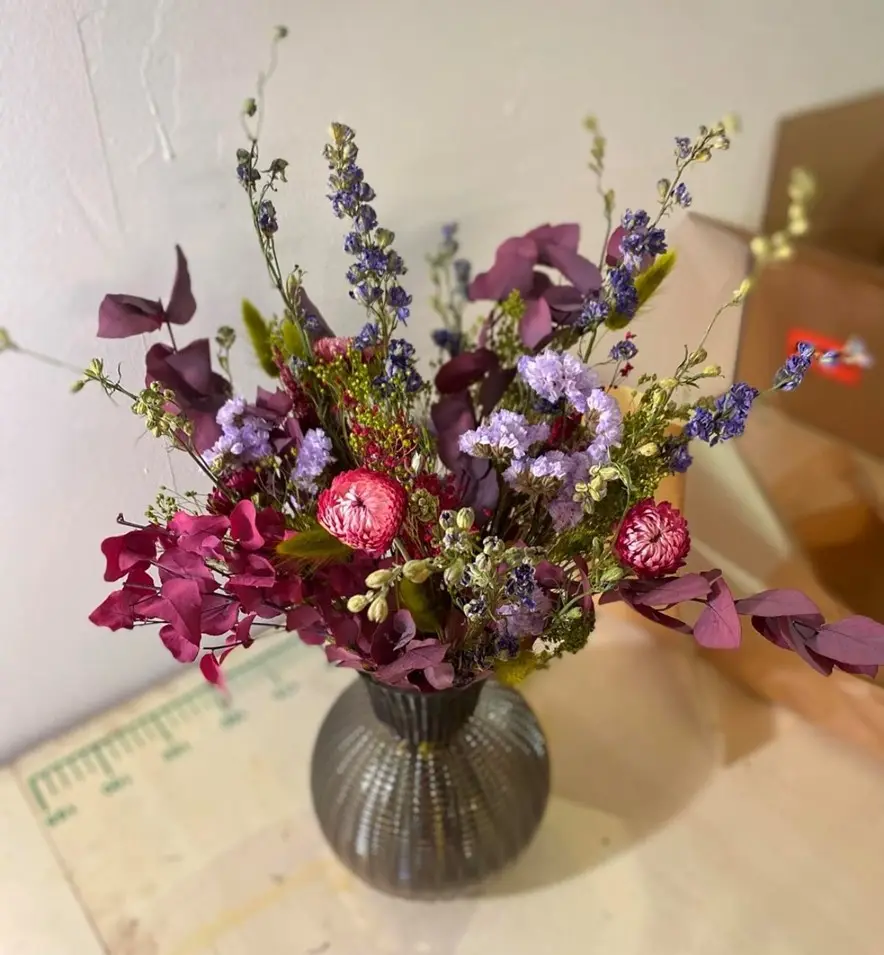 A vibrant, textured glass vase holds a dense arrangement of dried flowers including deep purple eucalyptus, pink and purple…