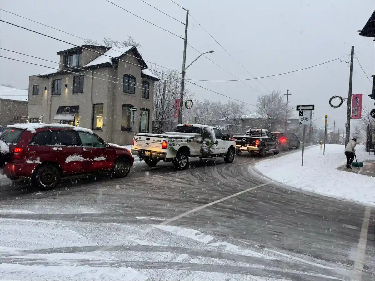 A snowy street scene with cars and a white pickup truck with 'ScpConcreteProsInc.com' branding. A person shovels snow on the…
