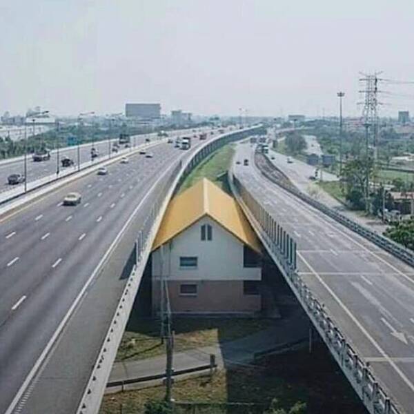 a house with a yellow roof sits directly under a multi-lane highway, with traffic flowing on both sides of the elevated…