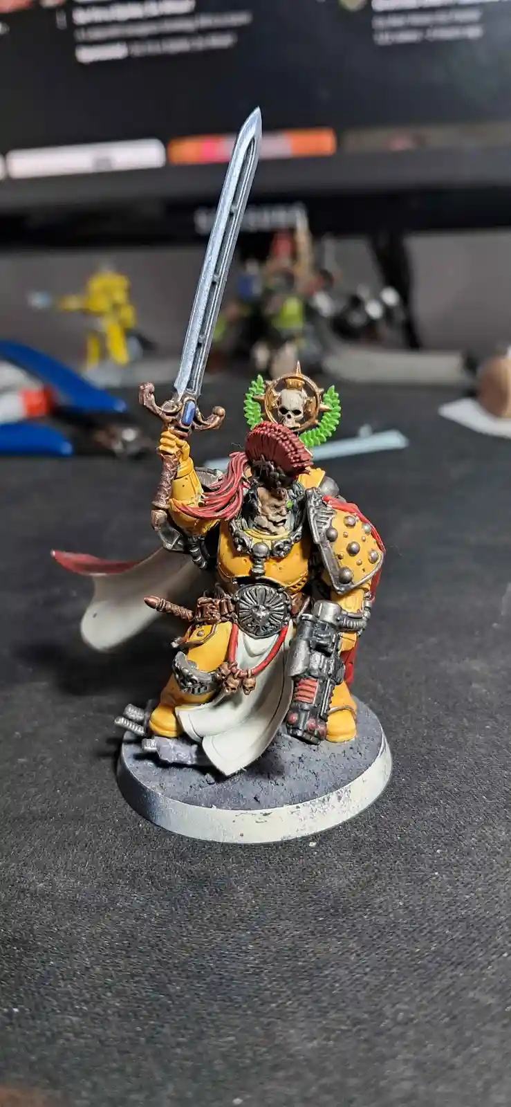 A painted miniature of a heavily armored, yellow-clad warrior with a skull-adorned helmet and a large sword raised high…