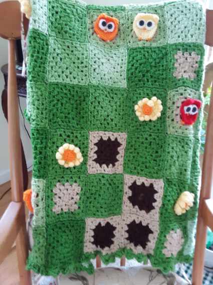 A handmade green crocheted patchwork blanket draped over a wooden chair, featuring various colorful appliqués including…