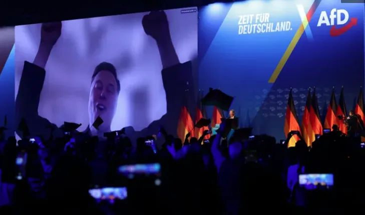 A large screen at a political rally shows a person with raised fists and an open mouth, while a crowd below holds up phones…