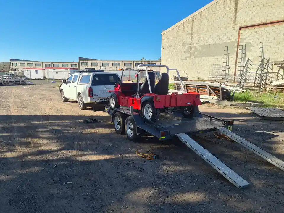 A white pickup truck is parked on a dirt lot, hitched to a flatbed trailer that carries a red, open-top, two-seater vehicle…