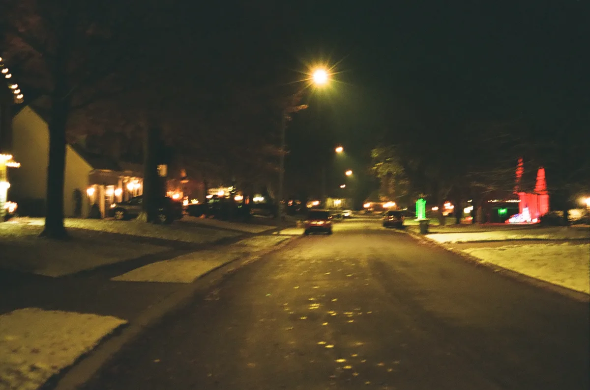 A blurry nighttime photo of a residential street with snow on the ground, streetlights, parked cars, and houses decorated…