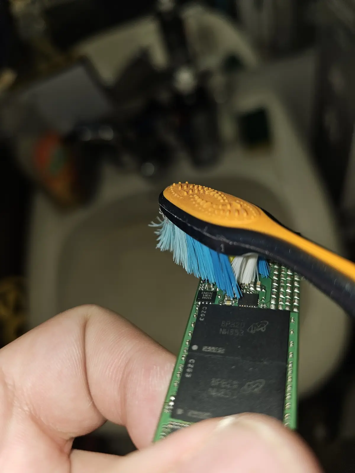 A close-up photo shows a person holding a small green circuit board with black chips, using a toothbrush with blue bristles…