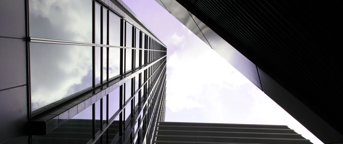 A low-angle photo shows two modern skyscrapers framing a bright sky, with one building's glass facade reflecting clouds…