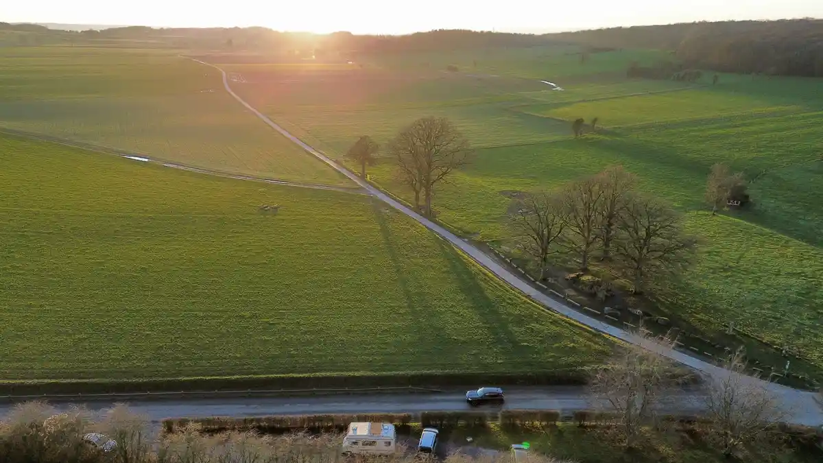 An aerial drone photo shows a winding rural road through green fields at sunset, with a black car driving along it and a…