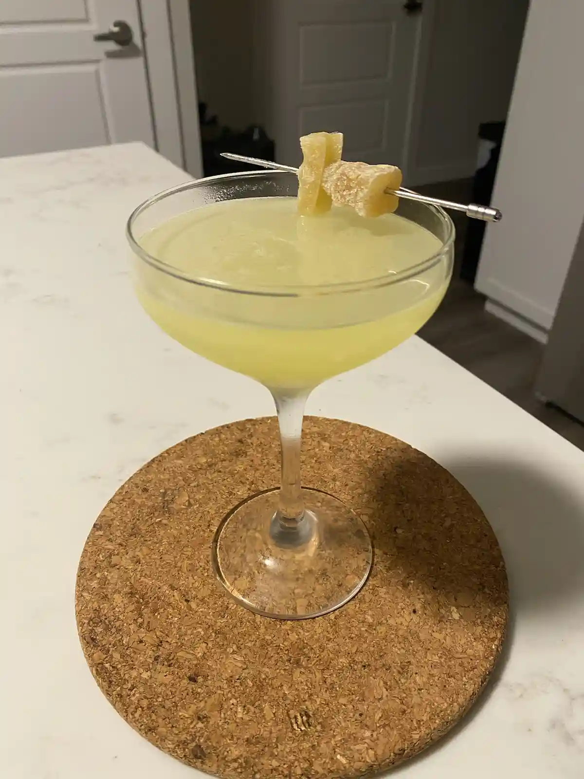A pale yellow cocktail in a coupe glass sits on a cork coaster atop a white countertop, garnished with two candied pineapple…