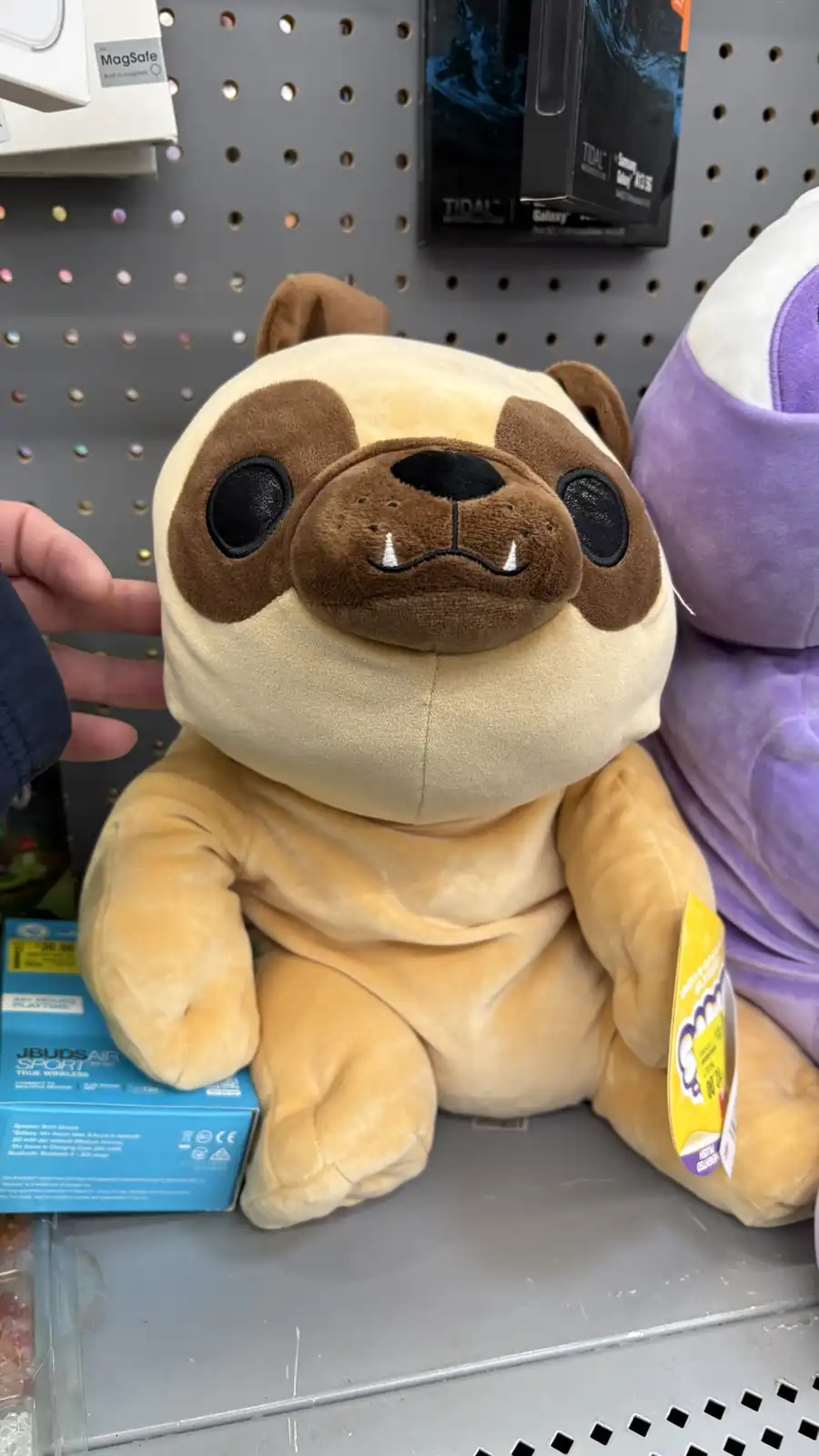 A tan plush pug toy with large black eyes and prominent white fangs sits on a store shelf, next to a purple plush toy and a…