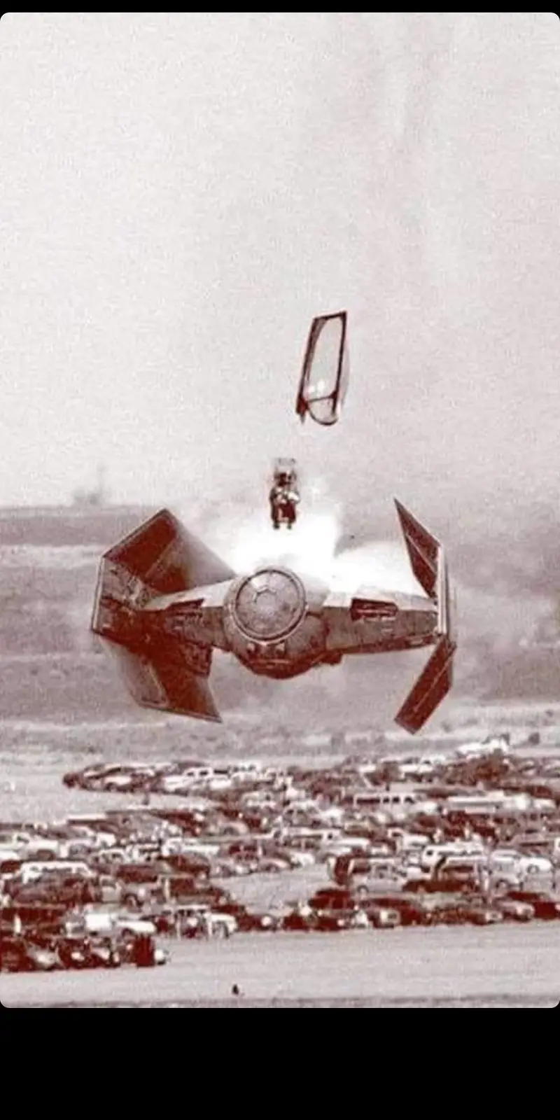 A sepia-toned image shows a Star Wars TIE fighter hovering over a crowded parking lot filled with cars. A small figure in a…