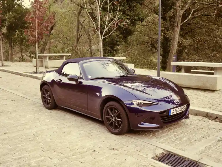 A dark purple Mazda MX-5 Miata convertible is parked on a cobblestone path in a park with trees and stone benches in the…