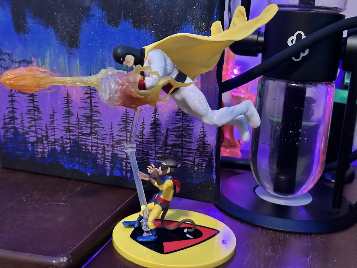 A Batman action figure with a yellow cape is posed mid-flight, leaving a fiery trail, while a smaller monkey figure in a…