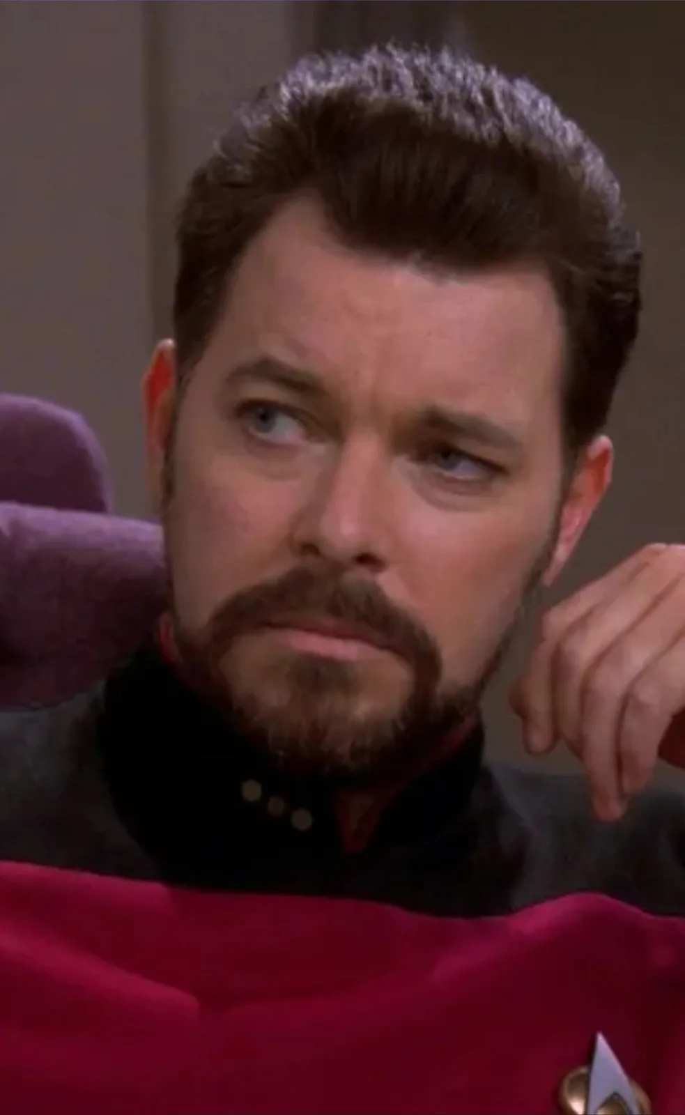A man with a beard and mustache, wearing a red Starfleet uniform with a gold insignia, looks sideways with a skeptical and…