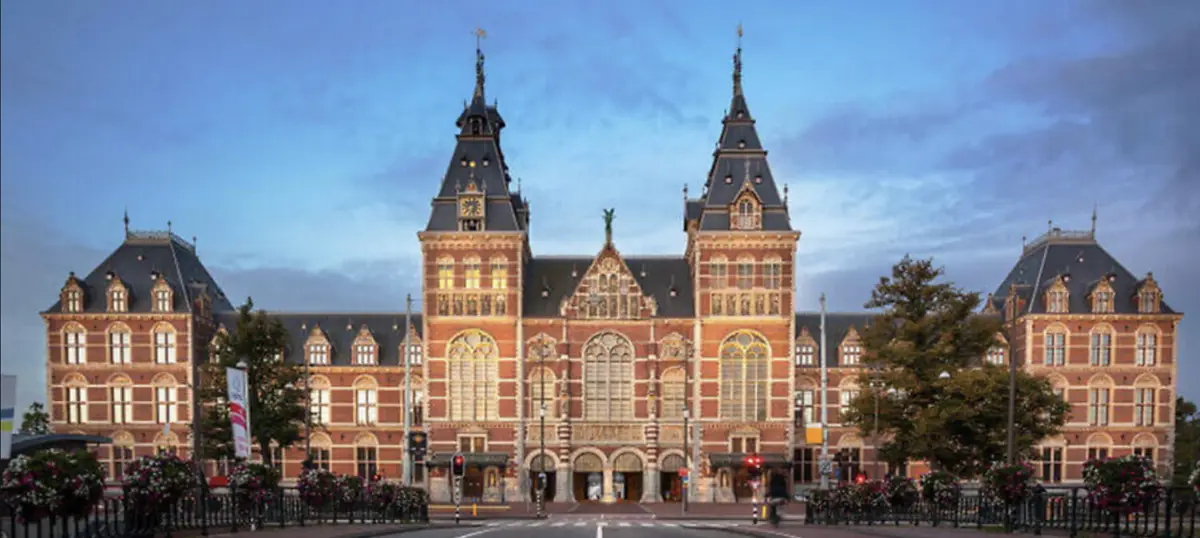 A wide shot of the grand, ornate, red-brick Rijksmuseum in Amsterdam, featuring two prominent towers and arched windows…
