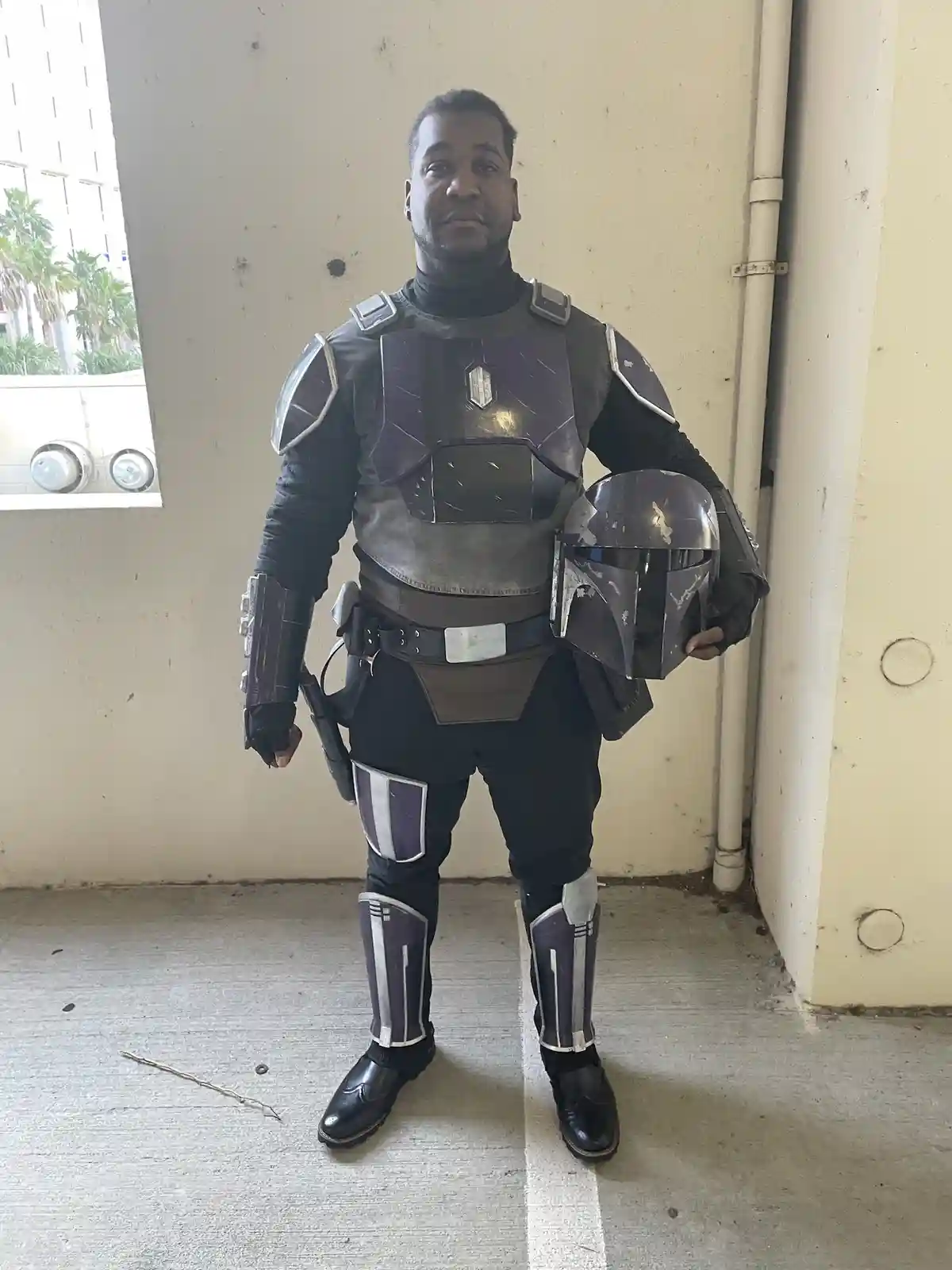 A man in a detailed Mandalorian armor costume, holding his helmet, stands in a parking garage. He is wearing black boots and…