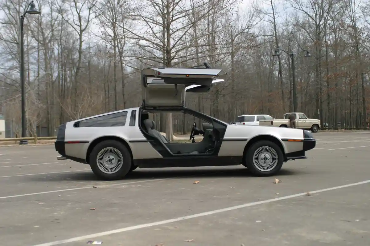 A silver DeLorean DMC-12 with its gull-wing doors and rear hatch open, parked in an empty lot surrounded by bare trees. The…