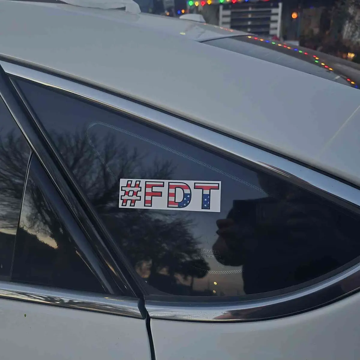 A close-up of a white car's rear side window featuring a rectangular sticker with the text '#FDT' in bold, block letters…
