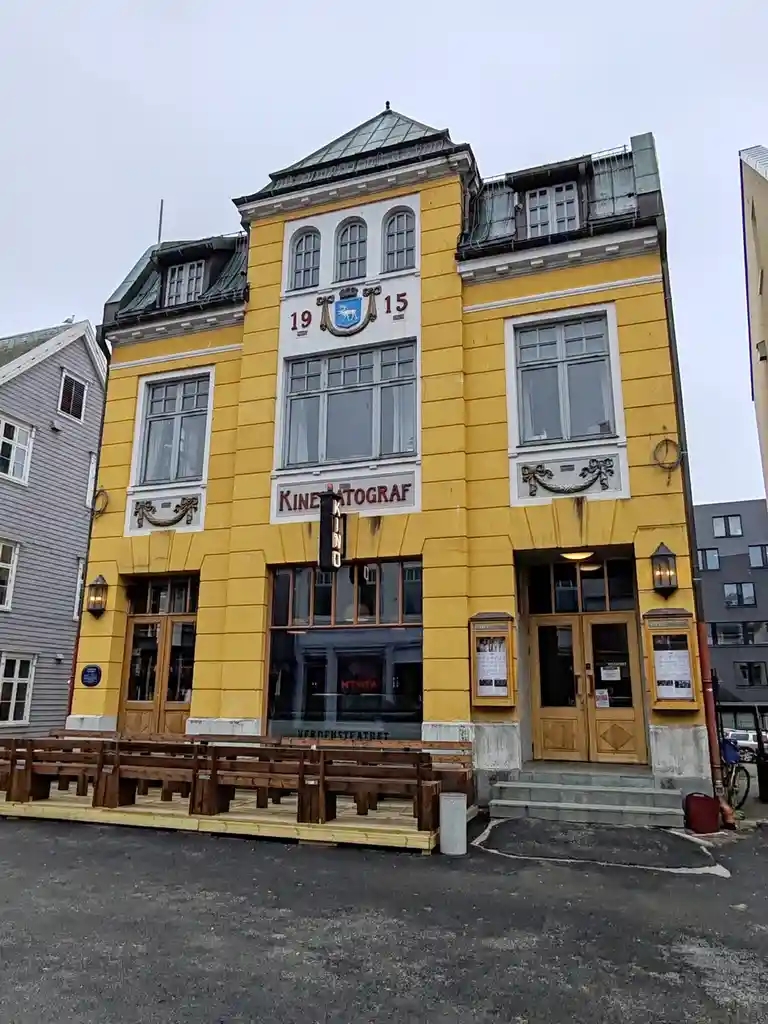 A yellow, three-story building with ornate white trim and a sign reading 'KINEATOGRAF' above a large window. The facade…
