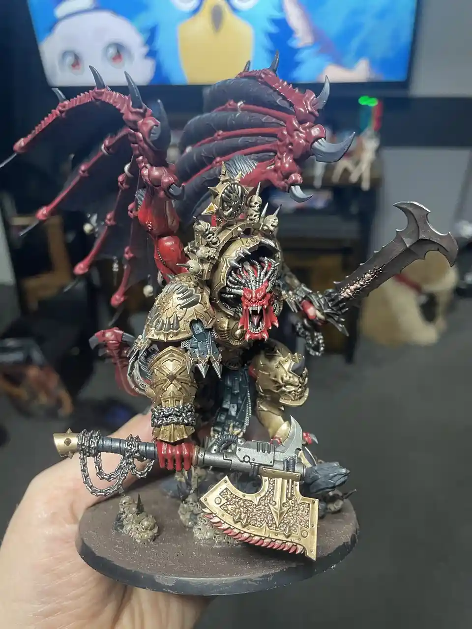 A hand holds a highly detailed, painted miniature of a demonic warlord with red and black wings, gold armor, and multiple…