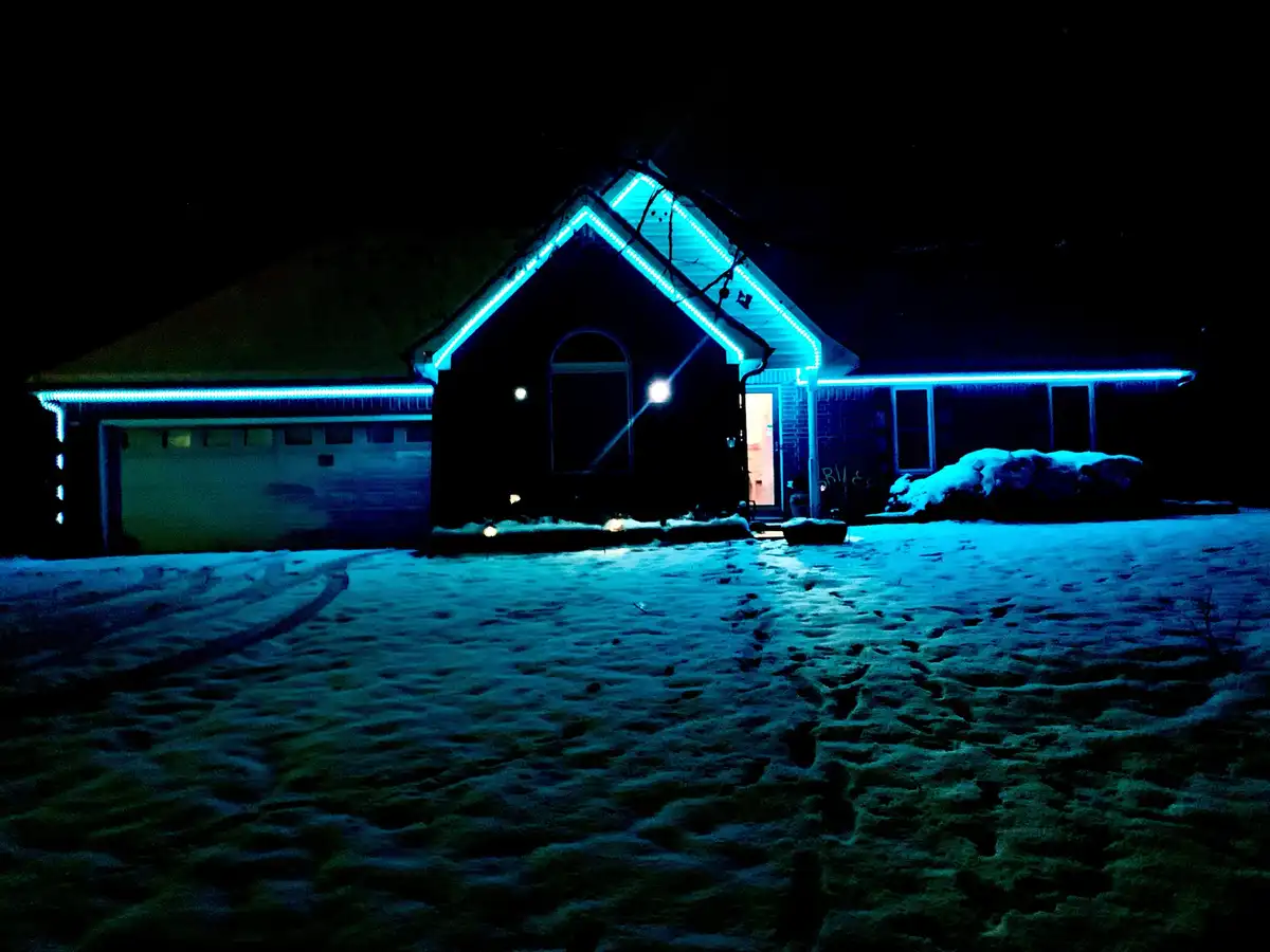 A house at night is illuminated by bright blue LED lights outlining its roofline, eaves, and windows, casting a blue glow on…