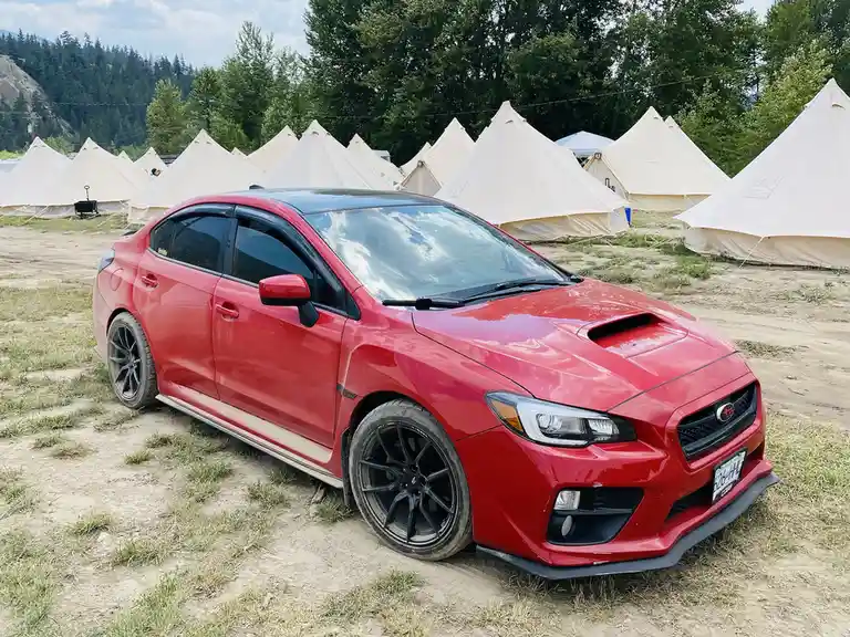 A red Subaru WRX STI is parked on dirt ground in front of a row of beige bell tents, with trees and hills in the background…