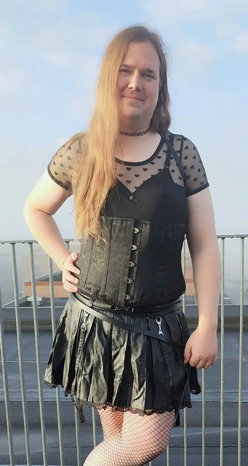 A person with long blonde hair stands on a rooftop wearing a black corset, a sheer heart-patterned top, a pleated skirt with…