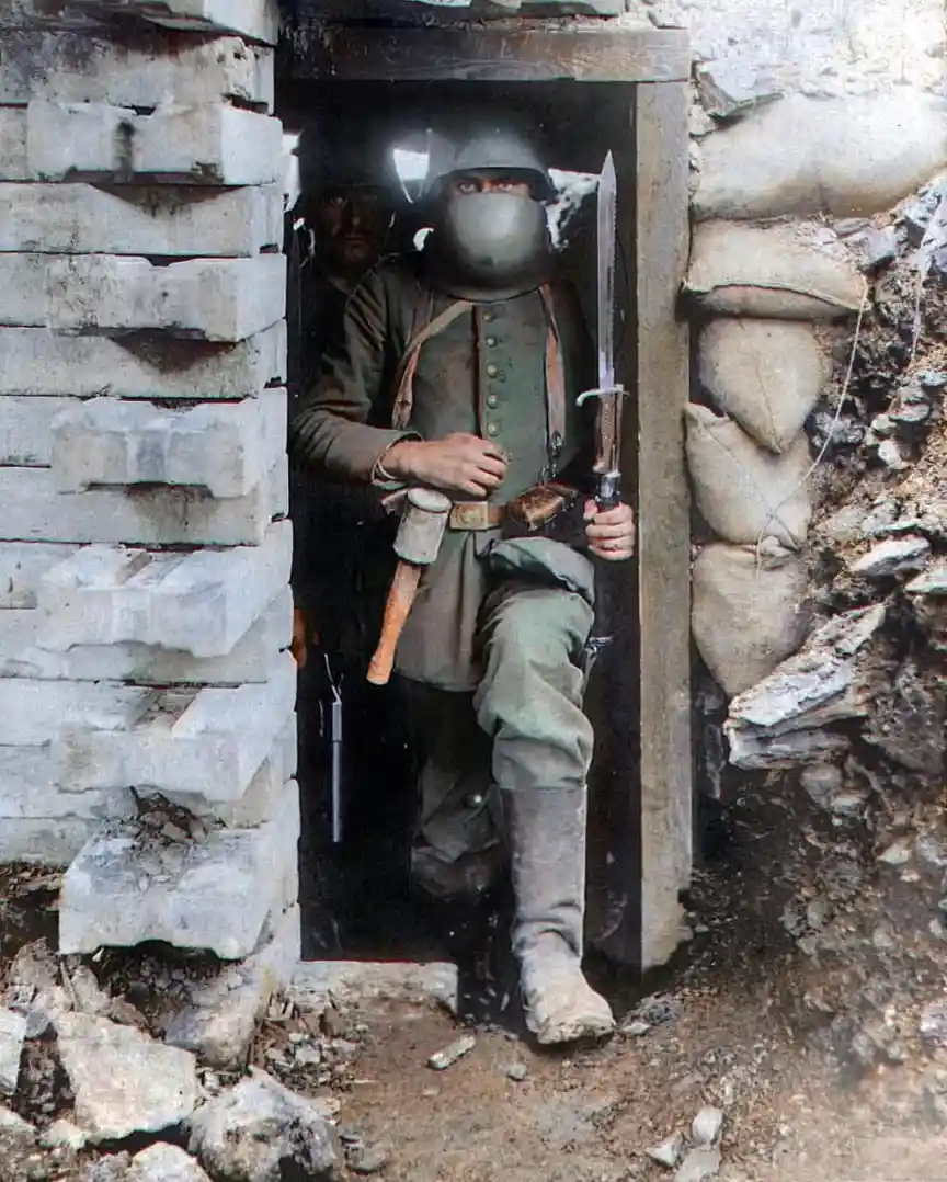A soldier in a trench, wearing a helmet and a full-face gas mask, holds a bayonet and looks out from a doorway made of…