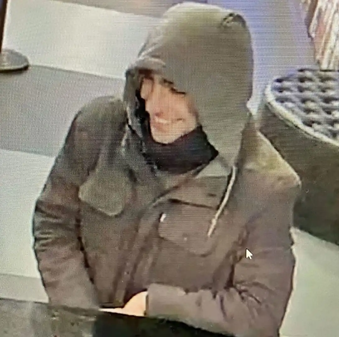 A grainy security camera image shows a person wearing a dark hooded jacket and a dark scarf, smiling broadly while seated at…