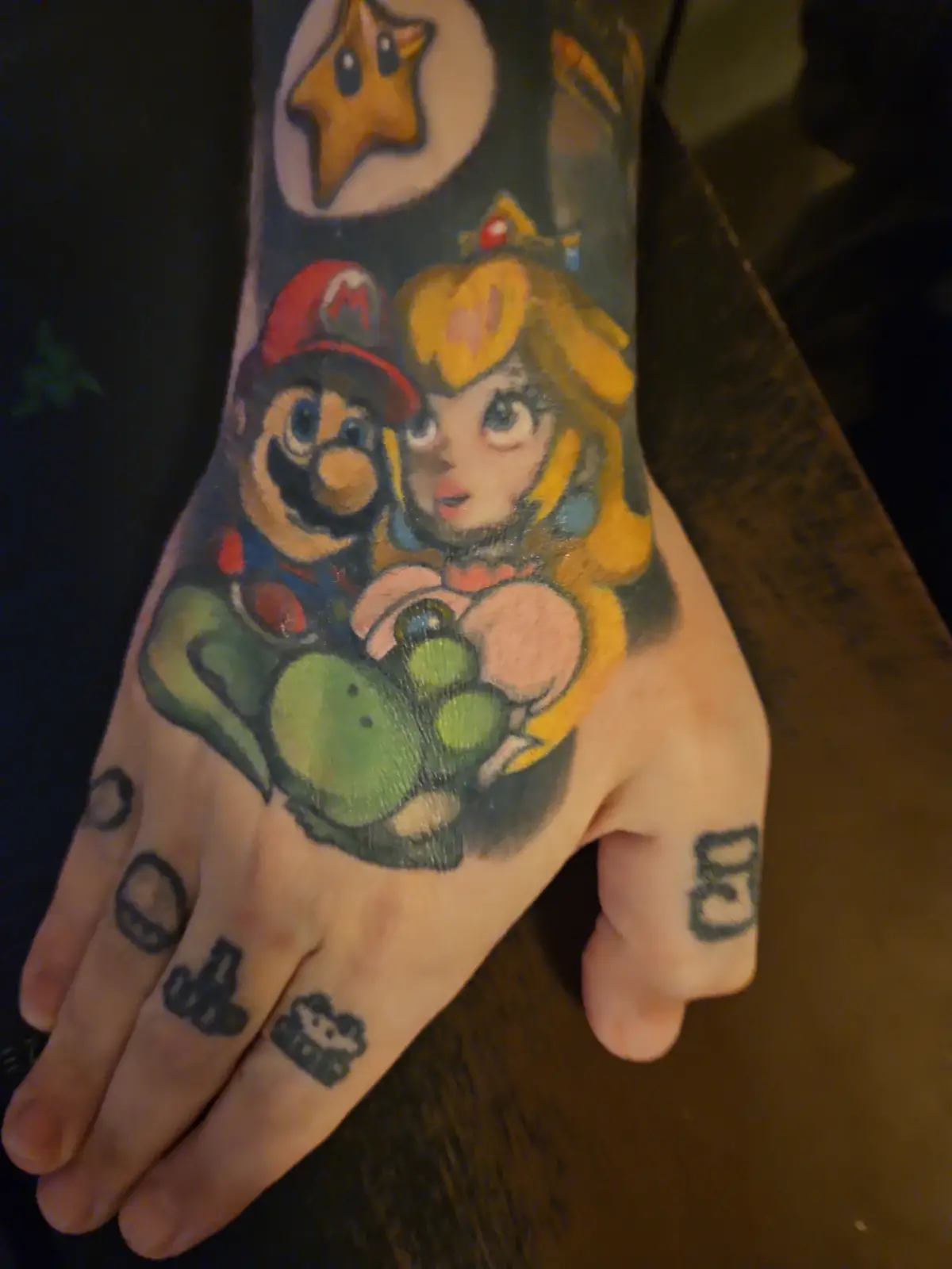 A hand with a colorful tattoo of Mario, Peach, and Yoshi on the palm and wrist, with a star above them. The fingers have…