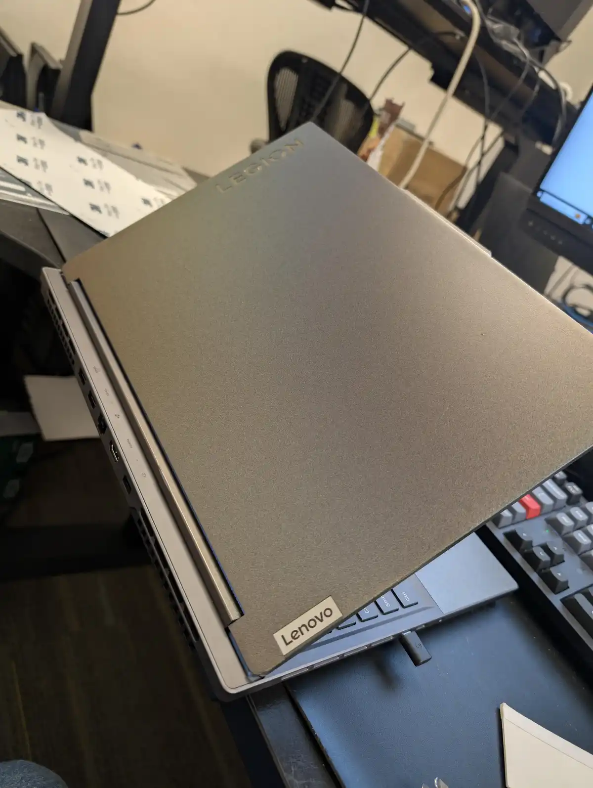 A closed gray Lenovo Legion laptop is shown at an angle on a desk, revealing its ports and the brand logo. The overlaid text…