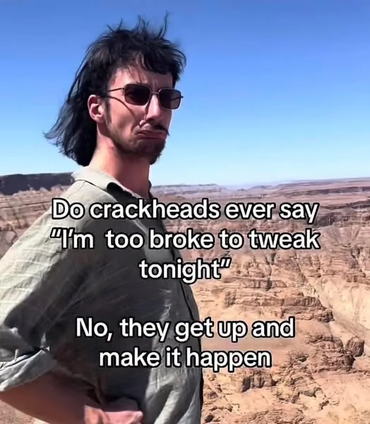 A man with dark hair, sunglasses, and a beard stands in a desert canyon with a serious, slightly smug expression. The…