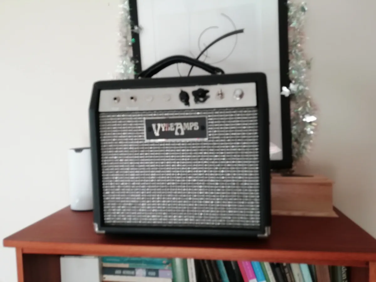 A black and silver 'Vyseamps' guitar amplifier sits on a wooden shelf, with a white smart speaker to its left and a framed…