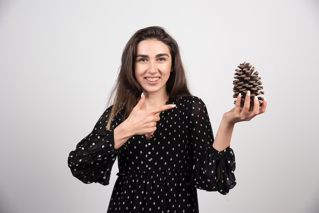 a smiling woman in a black polka-dot dress holds a pine cone in one hand and points at it with the other, against a plain…