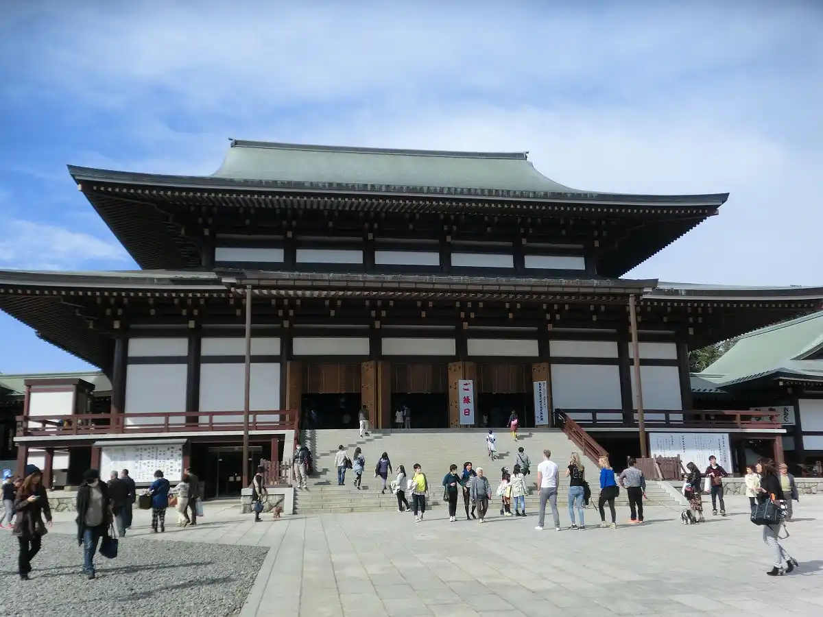 A wide-angle photo shows the grand, traditional Japanese temple structure with a large, sweeping green roof and wooden…