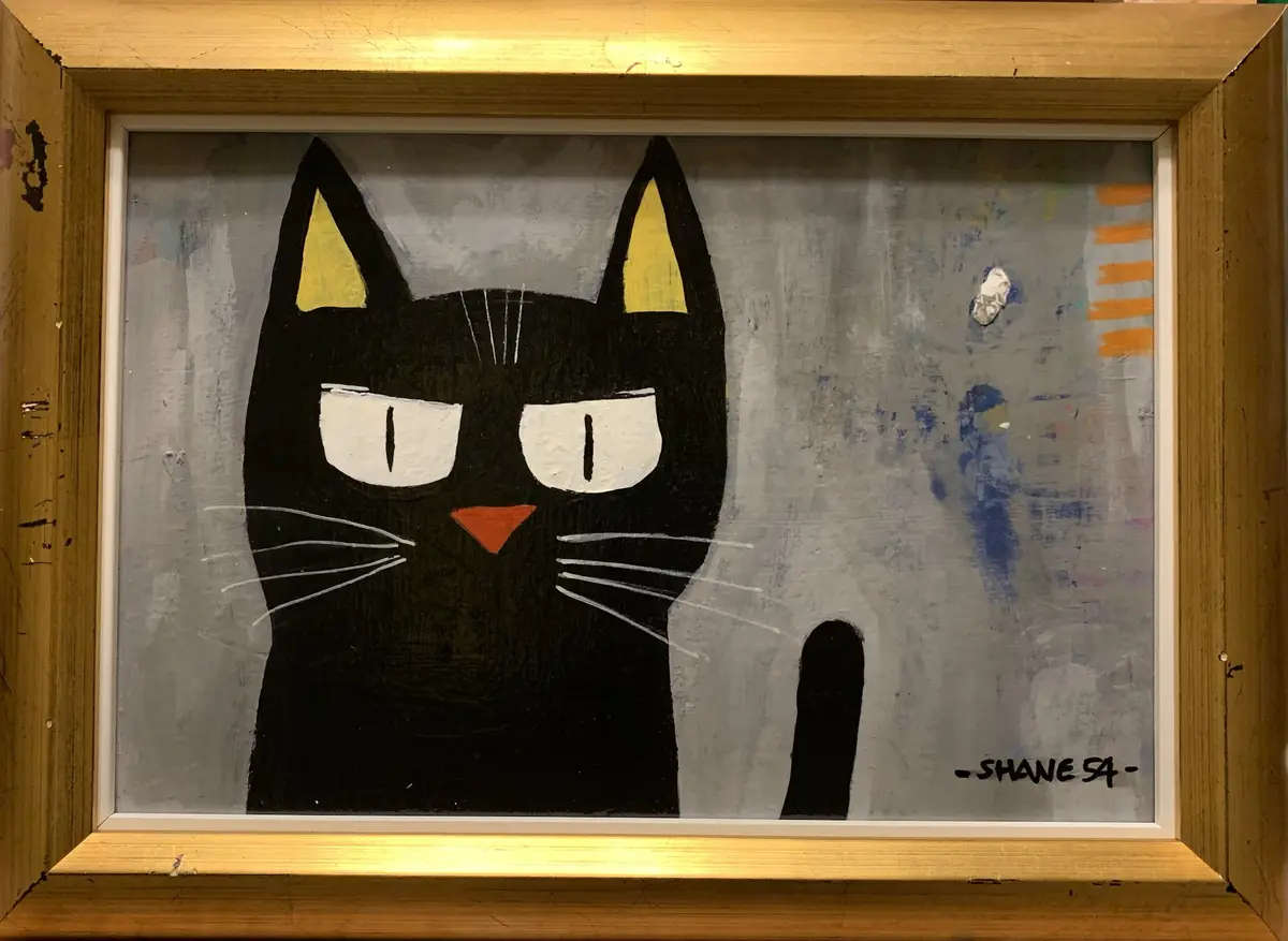 A framed painting of a stylized black cat with yellow inner ears, white eyes, and an orange nose, set against a gray…
