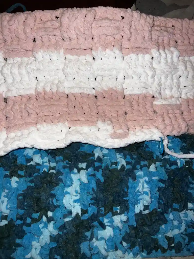 A close-up photo shows a thick, chunky knit blanket with horizontal stripes of pink, white, and a faded pink, resting on a…