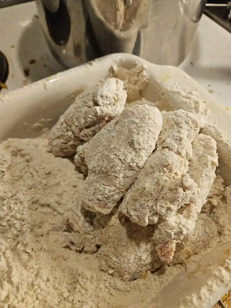 A close-up photo shows several raw chicken pieces heavily coated in flour, sitting in a white ceramic dish filled with the…