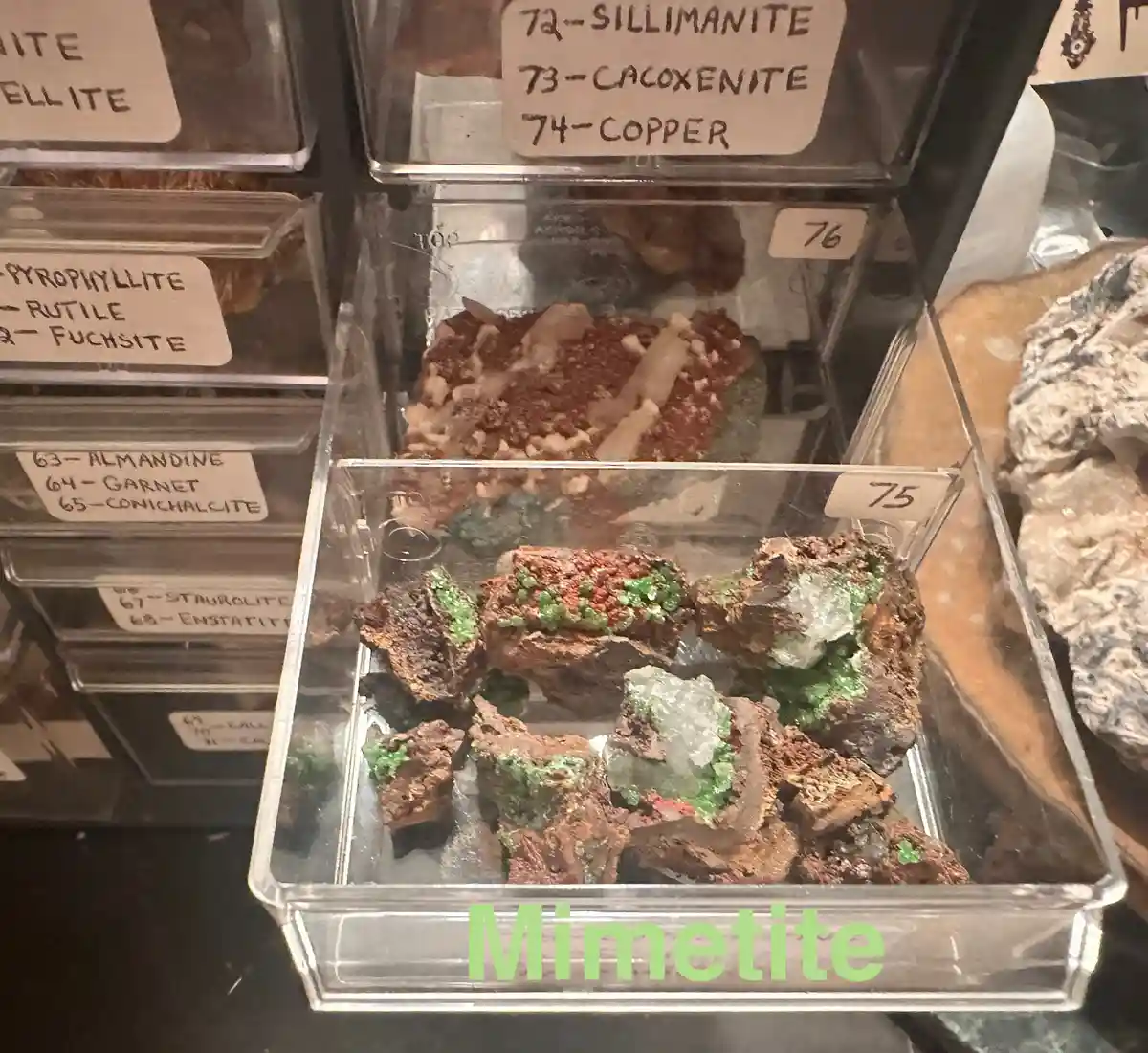 A close-up photo shows a clear plastic drawer labeled '75' containing rough, brownish mineral specimens with greenish…
