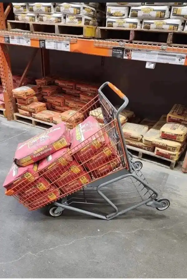 A shopping cart overflowing with red and beige bags of Quikrete concrete mix is tilted precariously on a concrete floor…