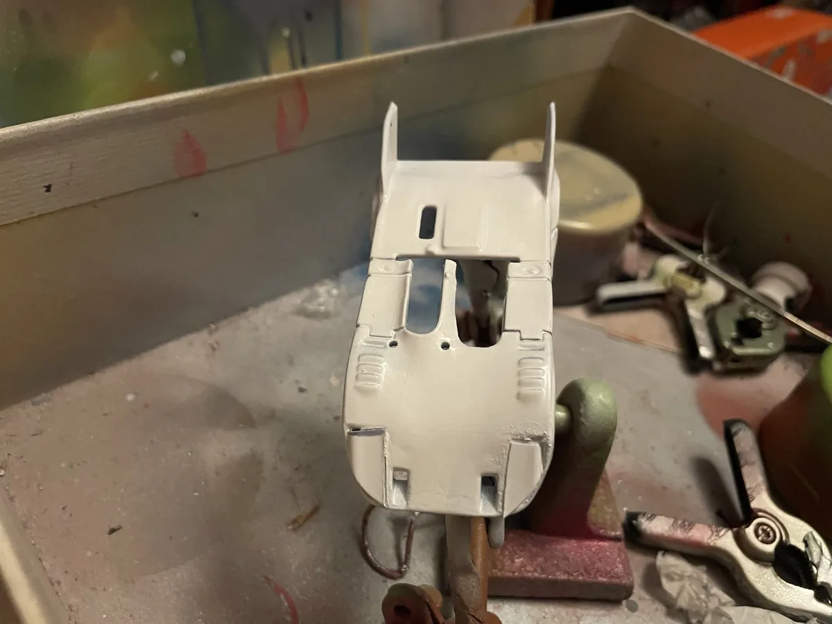 A white-painted model of the Batmobile is shown from the rear, mounted on a clamp in a cluttered workshop with tools and…
