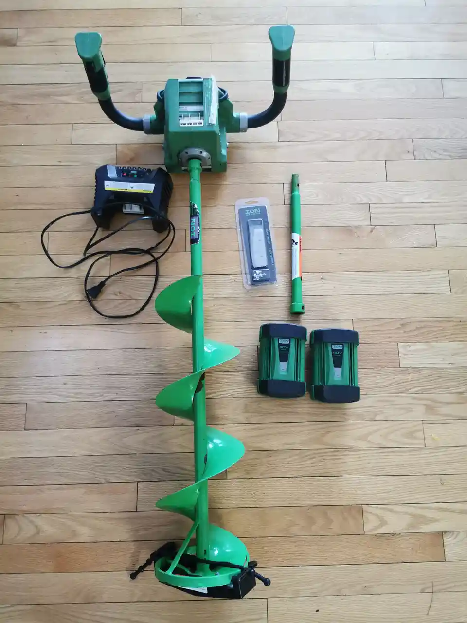 A green cordless ice auger and its accessories, including two batteries, a charger, and spare parts, are laid out on a light…