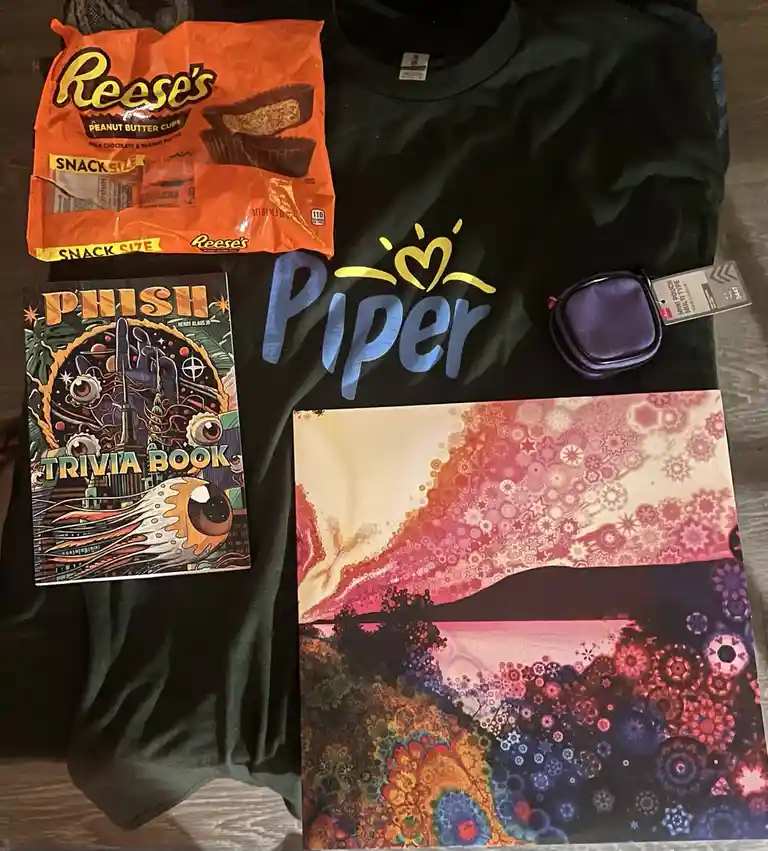 A flat-lay photo displays a black t-shirt with 'Piper' printed in blue and a yellow heart above it, alongside a bag of…