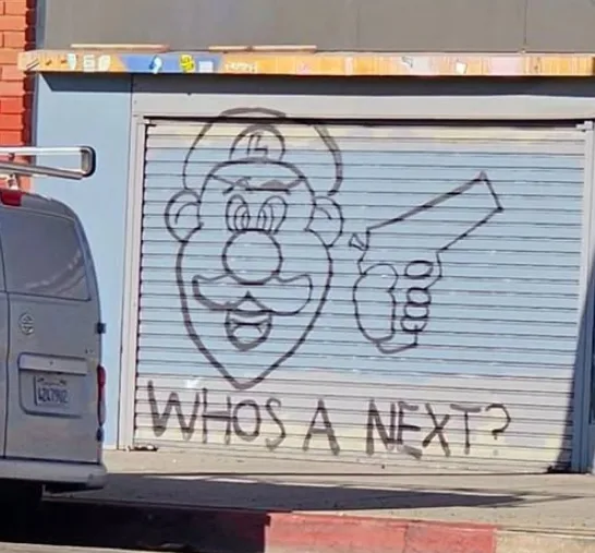 A light blue metal roll-up door has black graffiti of a cartoon character resembling Mario holding a gun, with the text…
